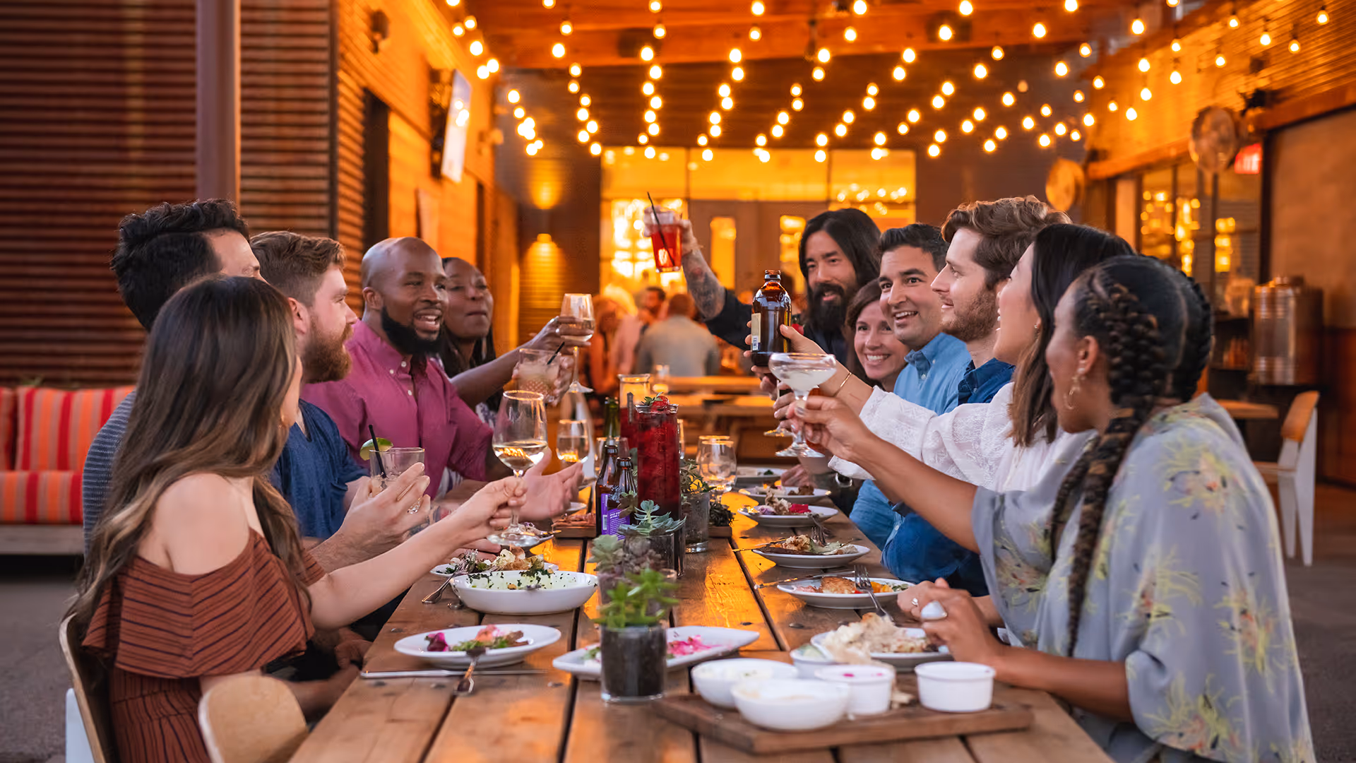 Group of friends enjoying an outdoor dinner party, raising glasses in a toast under string lights.